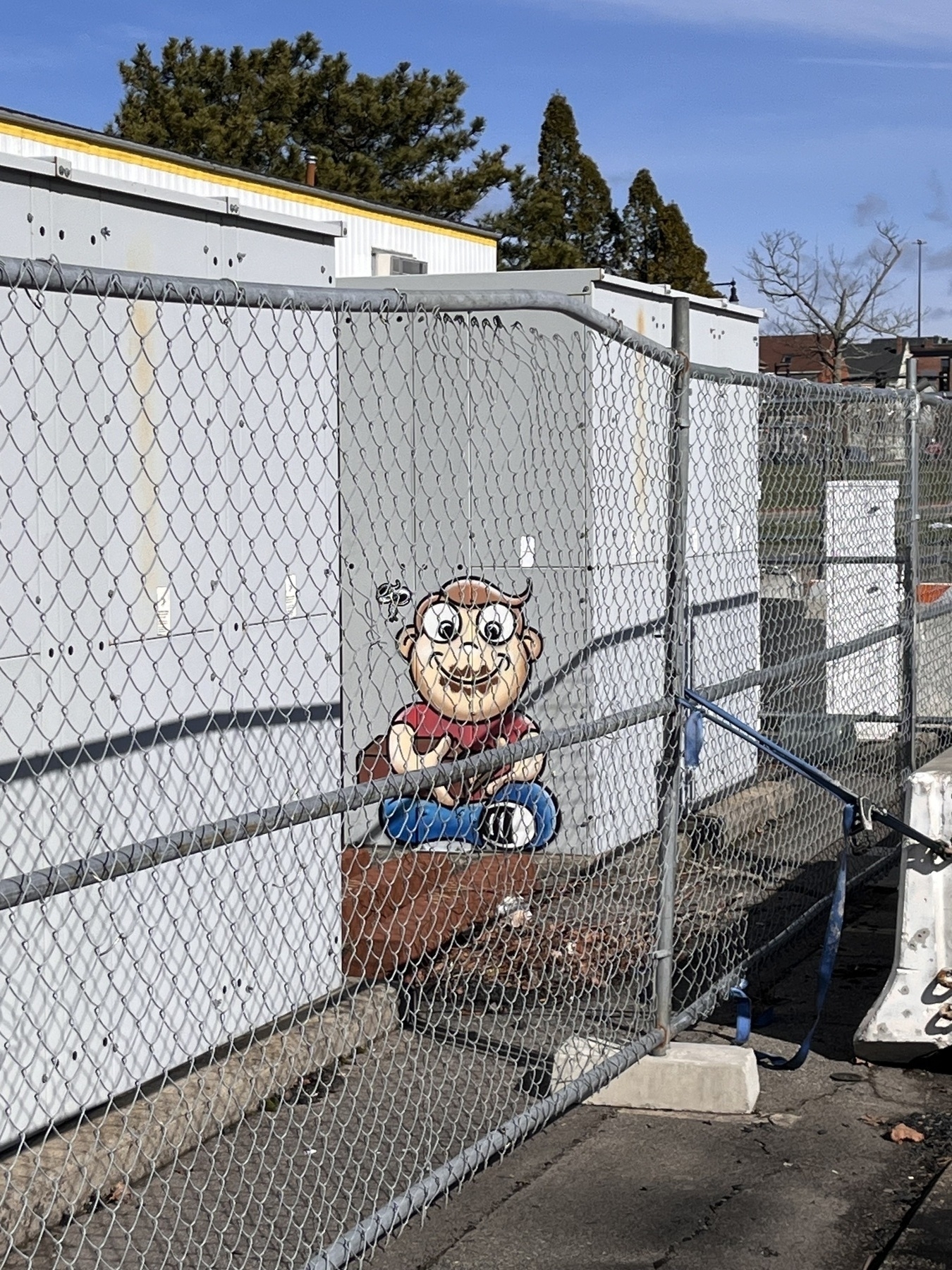 Graffiti on a large metal box behind a chain link fence of a cartoon boy sitting cross legged smeared in chocolate. He holds a big chocolate bar. 