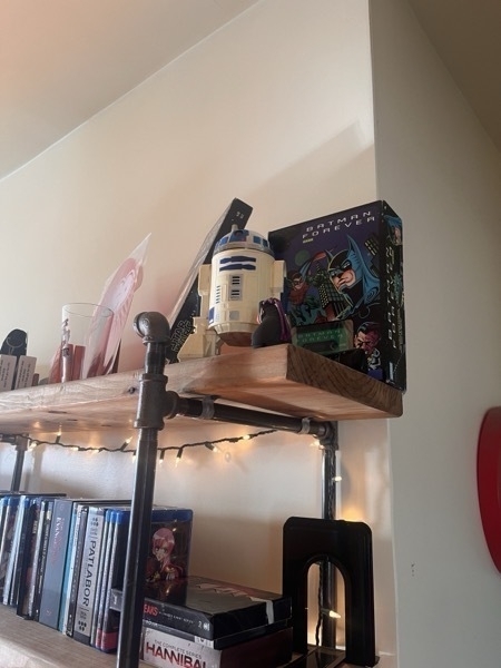 A shelf with an R2D2 figure on the top. Behind it is a Batman Forever board game propped against the wall. It is played with an audio cassette.