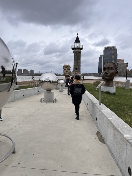 The Roosevelt Island lighthouse in the distance, with the path to it stretching in the foreground, against a stormy sky. Sculptures of Nellie Bly and reflective sphreres dot the path.