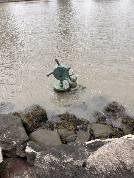 A cartoonish sculture just above the surface of the water. It's a coin with arms and legs, being dragged down by a little figure with the cash symbol for a face, coming out of a crotesque mouth. The mouth is mostly submerged in the water.