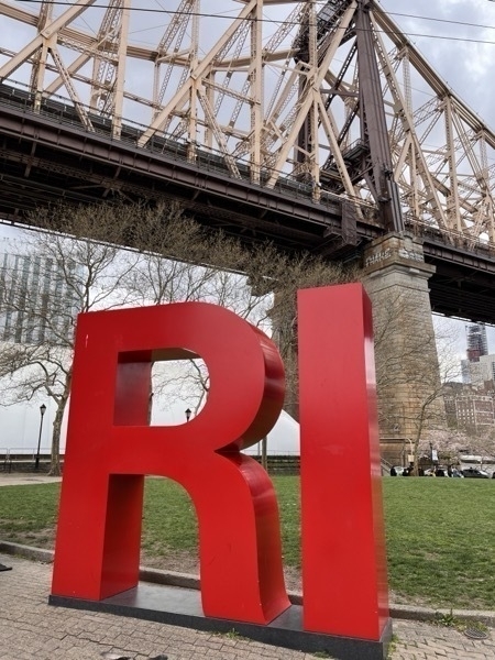 A sculpture of two large red letters, "RI", against a large bridge. The letters stand for Roosevelt Island.