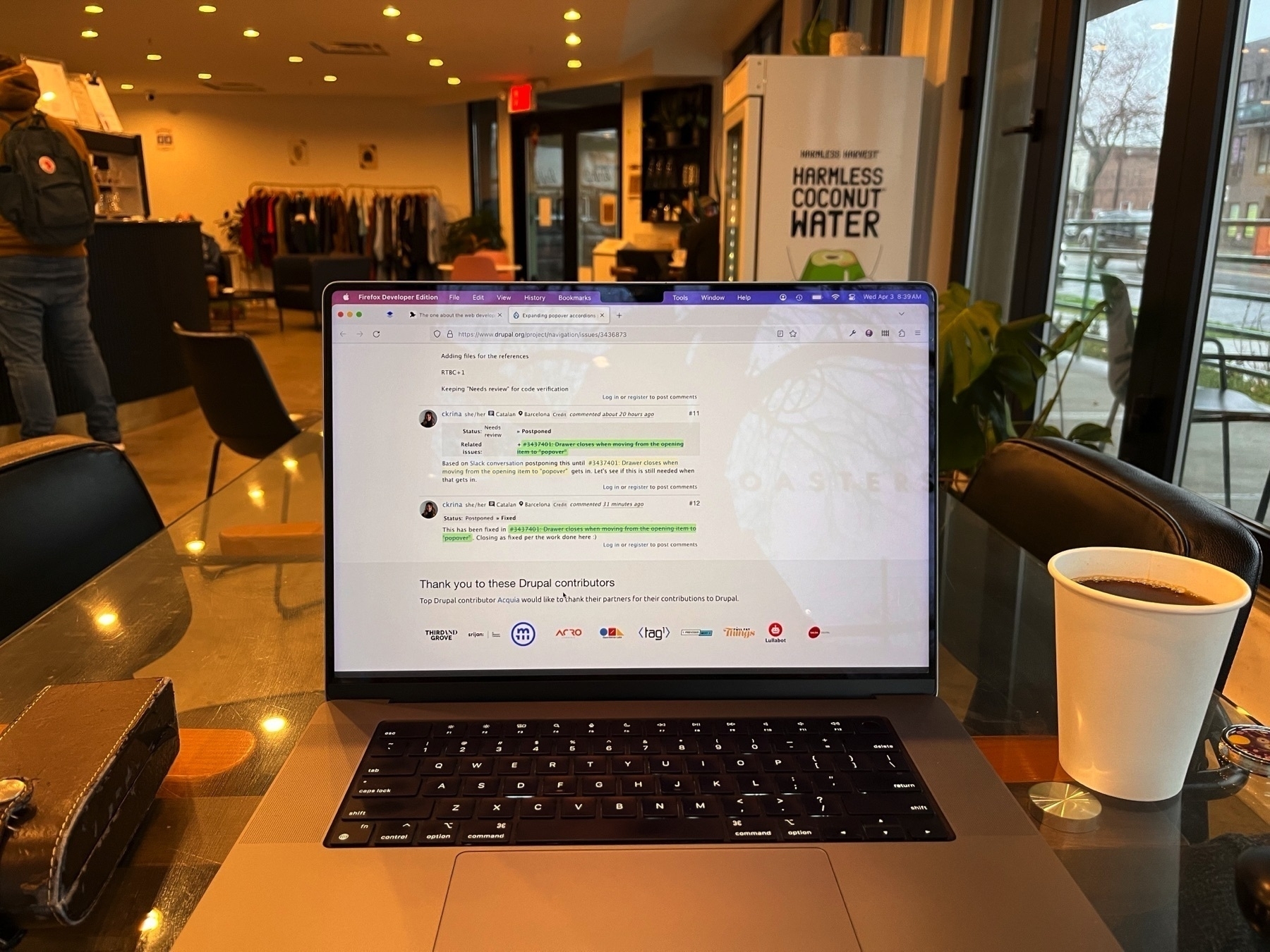 An open laptop on a glass table showing a Drupal navigation module ticket. There’s a hot coffee in a paper cup sitting next to it, and plants and coffee shop stuff in the background. There’s a fridge with “Harmless Coconut Water” branding on the side.