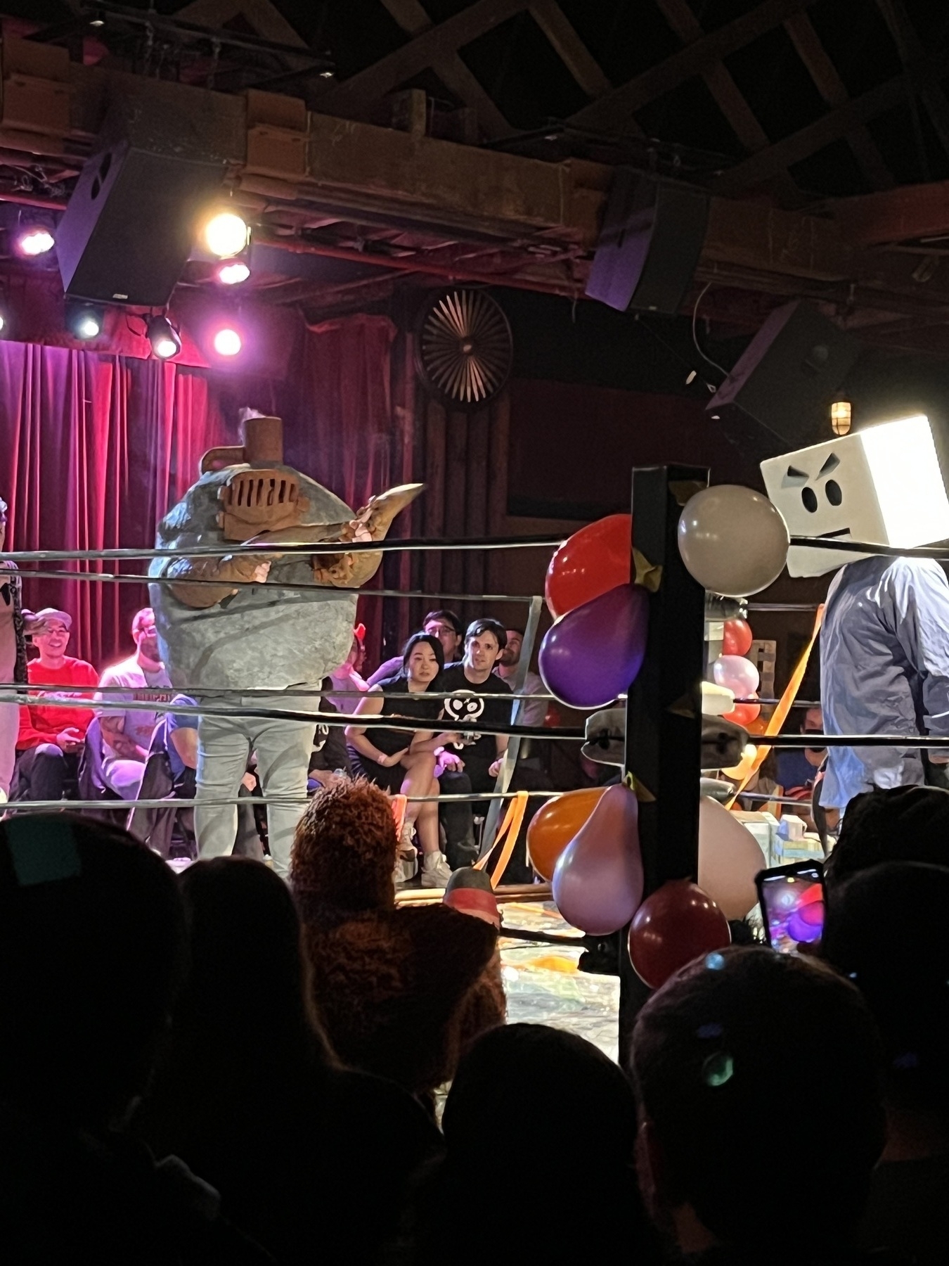 A wrestling ring with two people in costumes of giant monsters. One is wearing scrubs and a box on his head painted like a comic face, eyebrows downturned. The other is dressed as a boulder with two tentacles for arms. The boulder has a furnace grill for a face and a smokestack. 