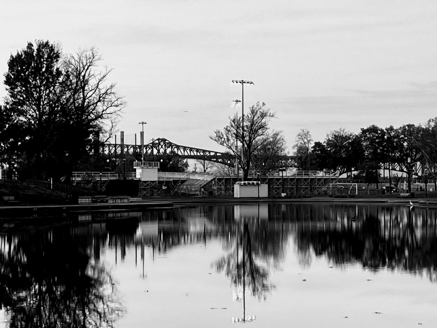 A moody black and white photo. A pond in the foreground, trees, a grandstand, and a metal suspension bridge behind. 
