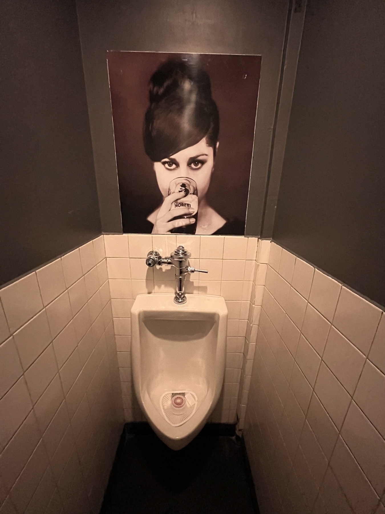A tiny room with a urinal at the end. A large photo of a women with a beehive hairdo drinking a beer and staring directly at you hangs above at eye level 
