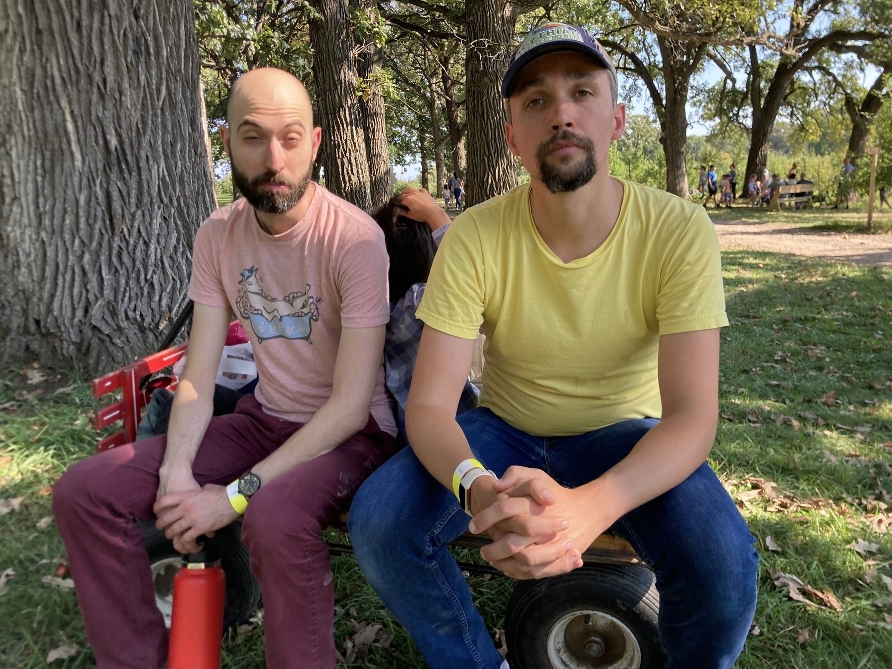Two white dudes sitting in a cart in a loose woods. The man on the right stares impassively into the lense. The man on the right looks like he smells a fart. 