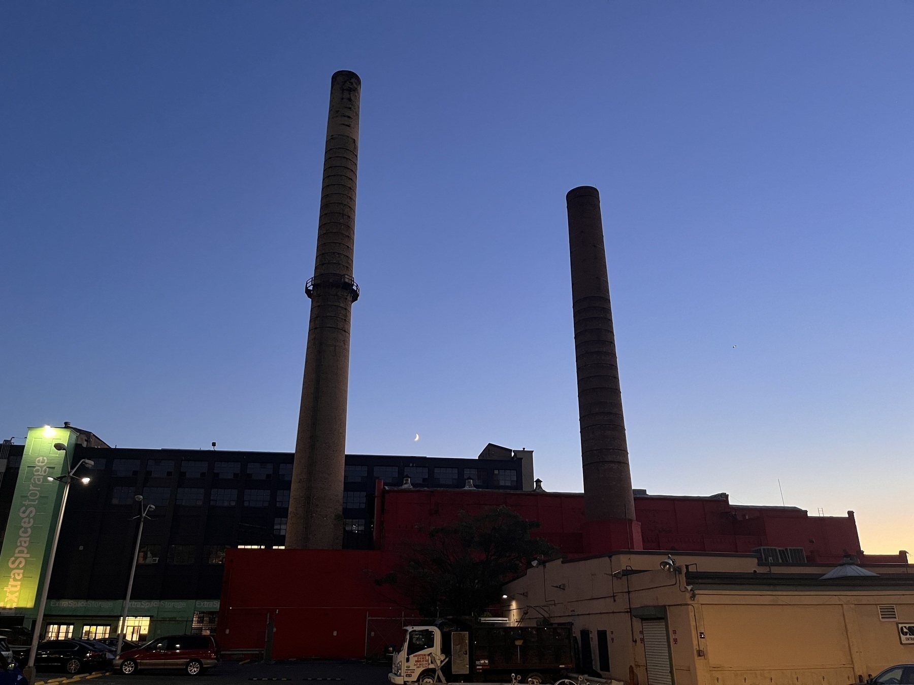 Two abandoned smoke stacks against a darkening sky, light still holding on in the corner. A sliver of a moon rests low between the smokestacks , right above industrial buildings. In the foreground is a truck. 