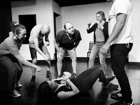  a monochrome photo of five improvisers on stage in various poses. One woman lays on the ground, a man stares intently off camera, others contort their bodies in approximations of objects. 