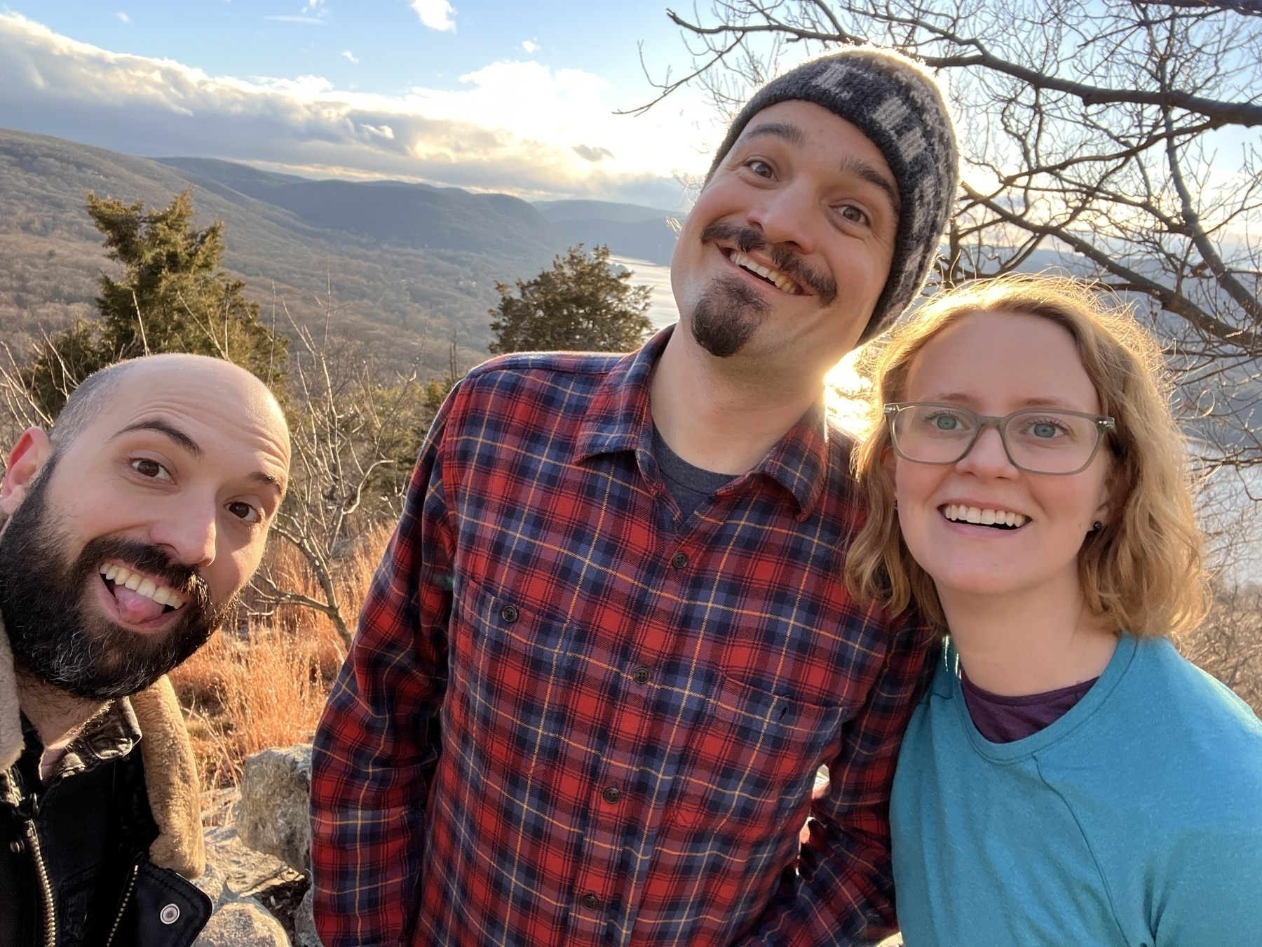 Three smiling white people, two men one women, in winter clothes with their backs to a sunset summit. 