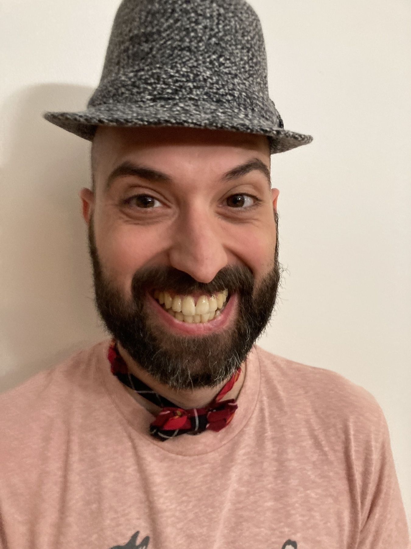 A white man grinning wide with a small fedora on the top of his head and a tiny bow tie. 