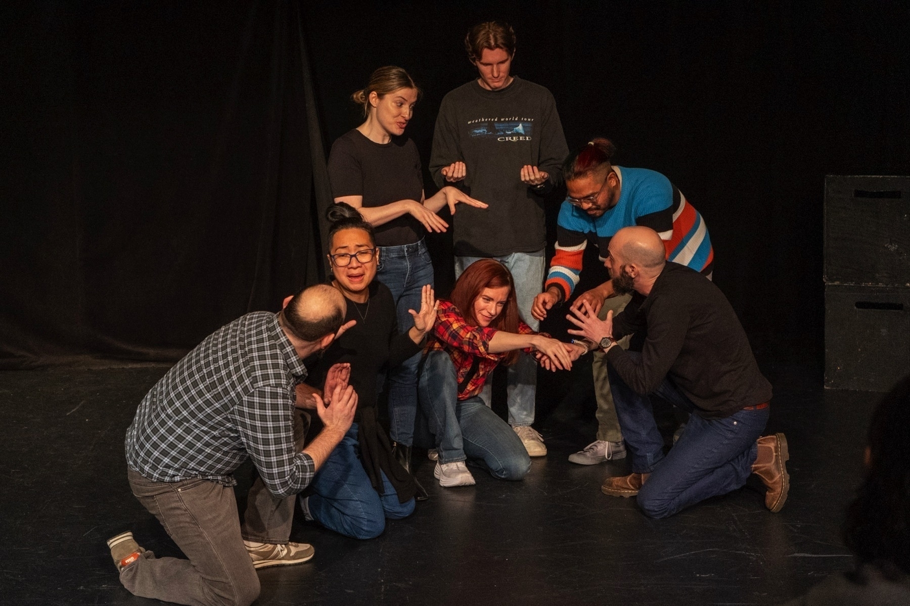 Seven individuals are actively participating in a dramatic scene on a black-box theater stage. They are gathered in a close semi-circle around a woman in the center who is crouched low to the ground, reaching out dramatically with one hand. The other performers are variously crouching or standing, with expressions and gestures of intense focus or concern directed towards the center. They are dressed in everyday casual wear, suggesting an improvisational or rehearsal setting. The stage backdrop is plain black, highlighting the actors, and the lighting casts well-defined shadows, enhancing the sense of a theatrical performance.