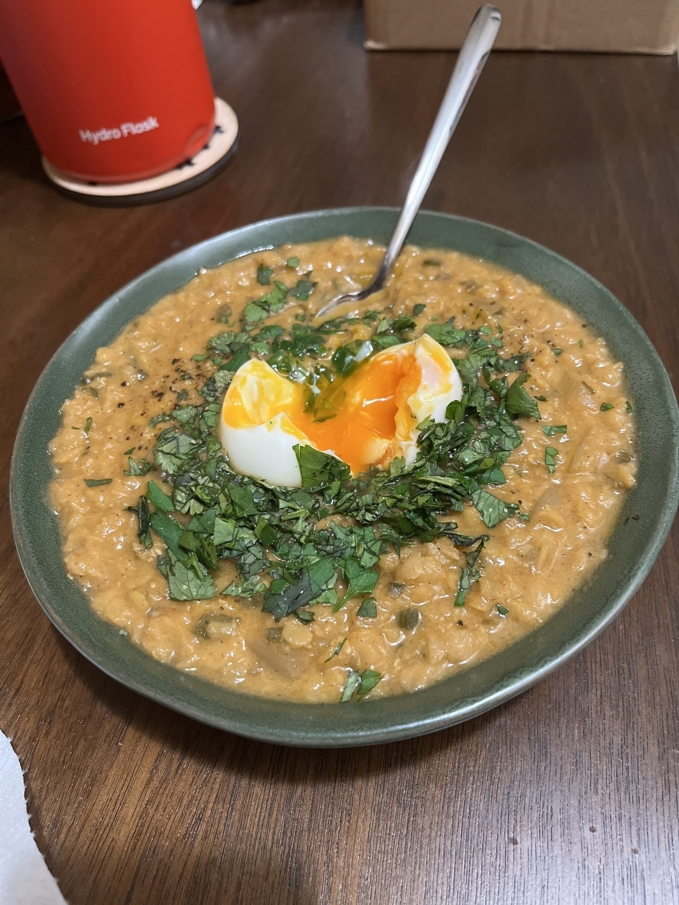 A green bowl with a yellow lentil curry inside. There’s chopped herbs on top, and an orange yolked soft boiled egg broken on top. 