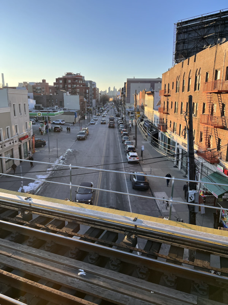 A clear day view from an elevated train platform looking down a bustling urban street lined with buildings. The street is filled with cars and a few pedestrians, with patches of snow on the sides and a clear blue sky above. The city skyline is faintly visible in the distance under the soft glow of the setting or rising sun.