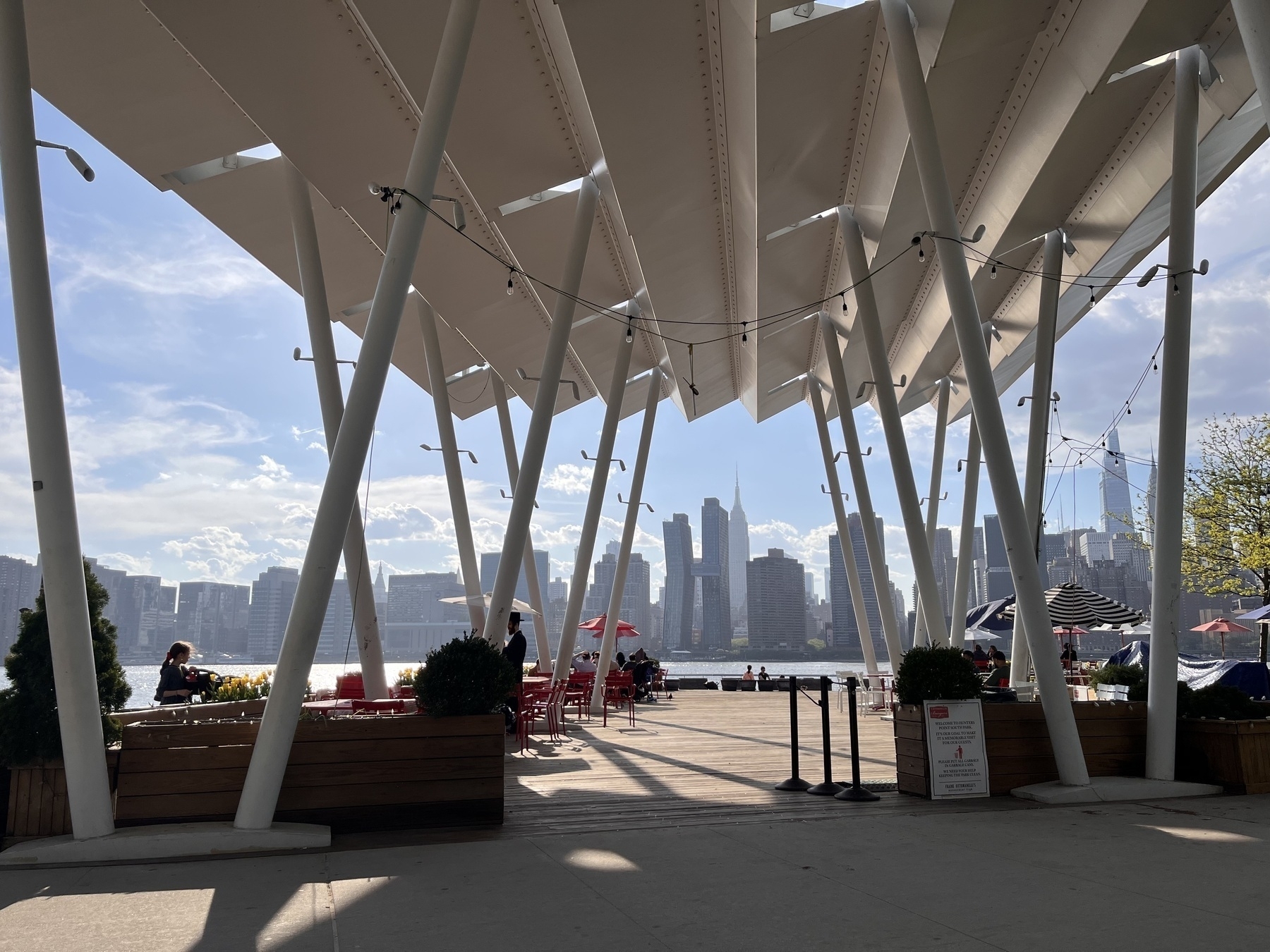 The New York city skyline over the water. There’s a big white serrated roof above the outdoor space. It’s a dock. There’s umbrellas. People kind of around. 