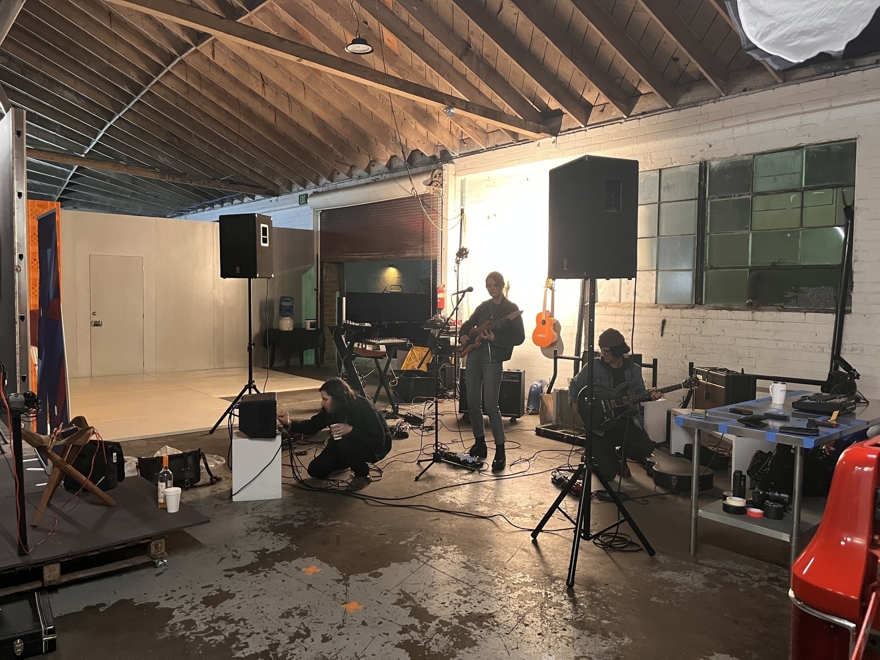 A industrial warehouse with three white people playing instruments. Two guitars and one guy crouching by a monitor. 