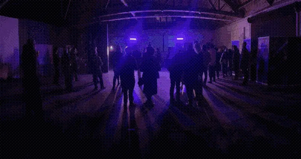 A dithered, dark shot of a small crowd in a warehouse. The people are staring towards purple lights.