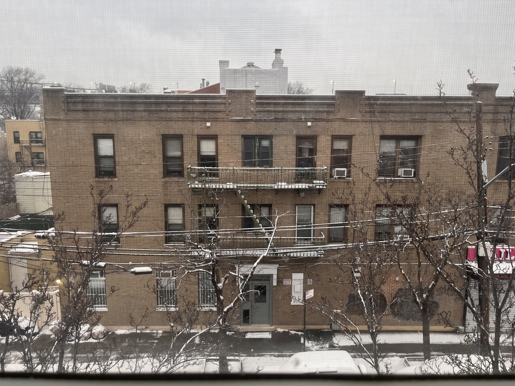 A three story brown stone building seen from a window on the third floor of the opposite building. A light dusting of snow coats everything. It’s especially visible on the fire escape. The sky is steel grey.  