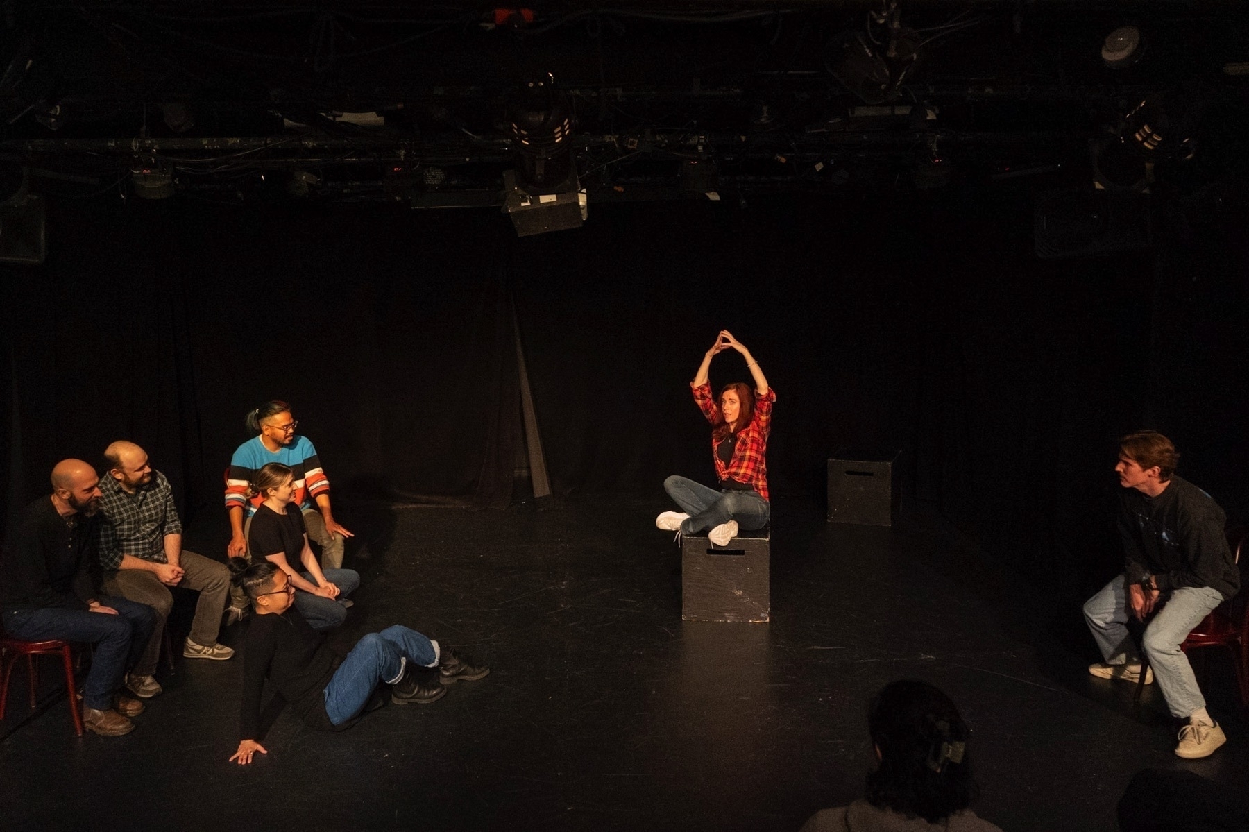 On a sparsely decorated stage, a group of seven people appears to be engaged in a theater workshop or improvisation scene. A woman in a red plaid shirt is perched on a black box, forming a peak with her hands above her head, possibly mimicking a mountain or roof. To her left, a man is squatting with his hands on his thighs, looking towards her. On the left side of the image, five people are seated or lying on the stage, with some looking toward the woman on the box. They seem to be attentive and possibly reacting to her action. The stage is simply set with a black backdrop and stage lighting equipment visible overhead. The audience perspective is from the back of the room, with the back of an audience member's head in the foreground.
