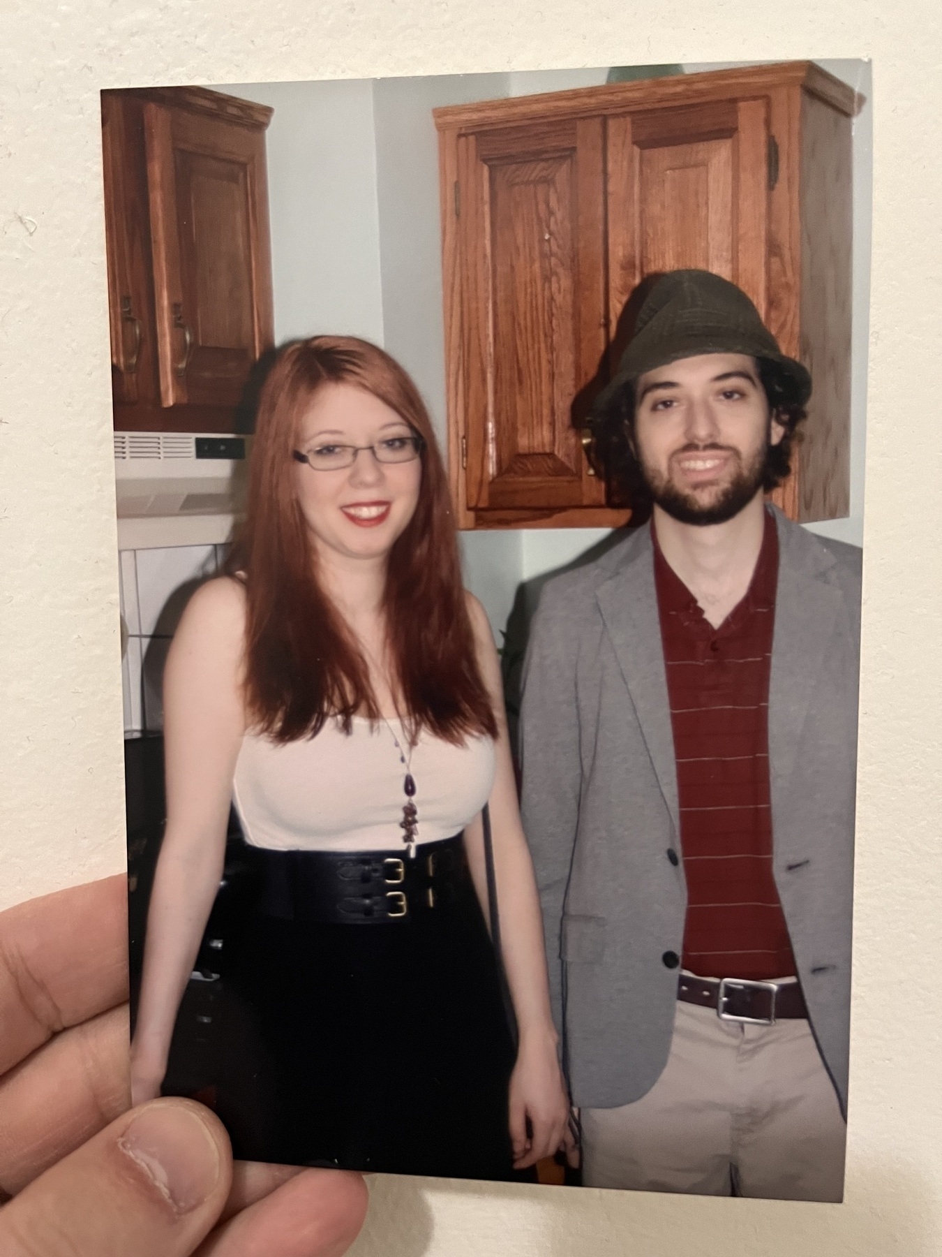Two young white peope standing awkwardly next to each other. The woman on the left, my sister, has long red hair and smiles somewhat uneasily. In the right, is me. I have a red shirt that doesn’t quite fit, khaki pants, a grey blazer, and on my head is a floppy, misshapen dark green fedora. I have a thin, untrustworthy beard. Unkept curls sprout from under the hat. My expression is haunted. We are in a kitchen.