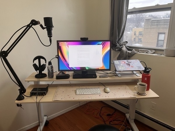 A desk caddy corner in a bare walled room. The desk has two levels, one for the monitor, and another for the keyboard. It is light, processed wood. On the left is a mic arm and audio deck on the lower level, and headphones and canned air on the upper level. In the middle is a 26 inch acer monitor on the upper level, and an old magic keyboard and even older wired mouse on the lower level. The lower level also sports an abstract, colorful star patterned mouse mat. On the right is a mac book pro sitting on top of a laptop stand, clamshell closed. There are various dumb stickers all over it. A mug of coffee and a water thermos sit on the far right on the lower level.