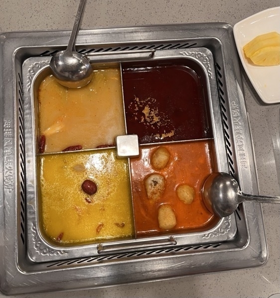 Four soup bases in the center of a table, two ladles sticking out of the broth. Some peppers and crispy tofu peak out. There's a small plate of pineapple next to the soup.