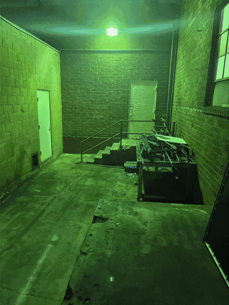 A green, dithered photo of a shallow allyway. There are brick buildings forming the corridor, with a cemement staircase leading to an embedded metal door. There's a pile of scrap metal neatly stacked in front of the staircase.