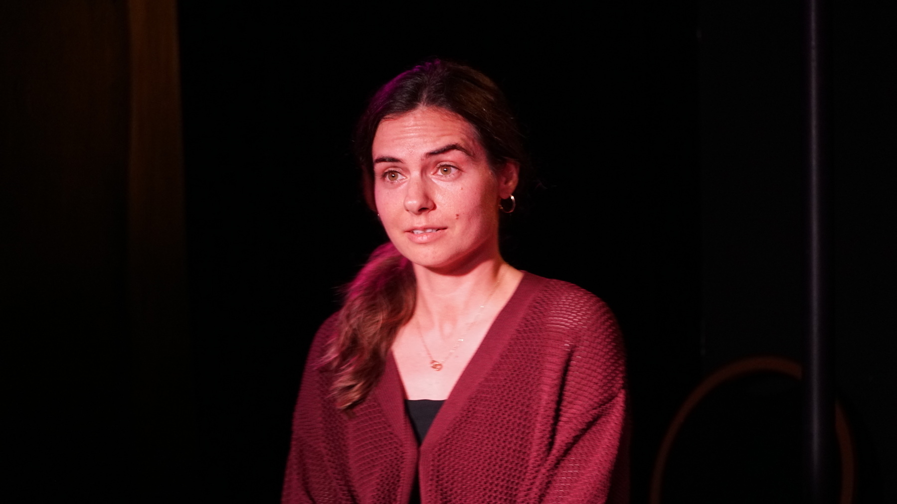 Auto-generated description: A woman with long hair, wearing a maroon sweater, is lit by a spotlight against a dark background.