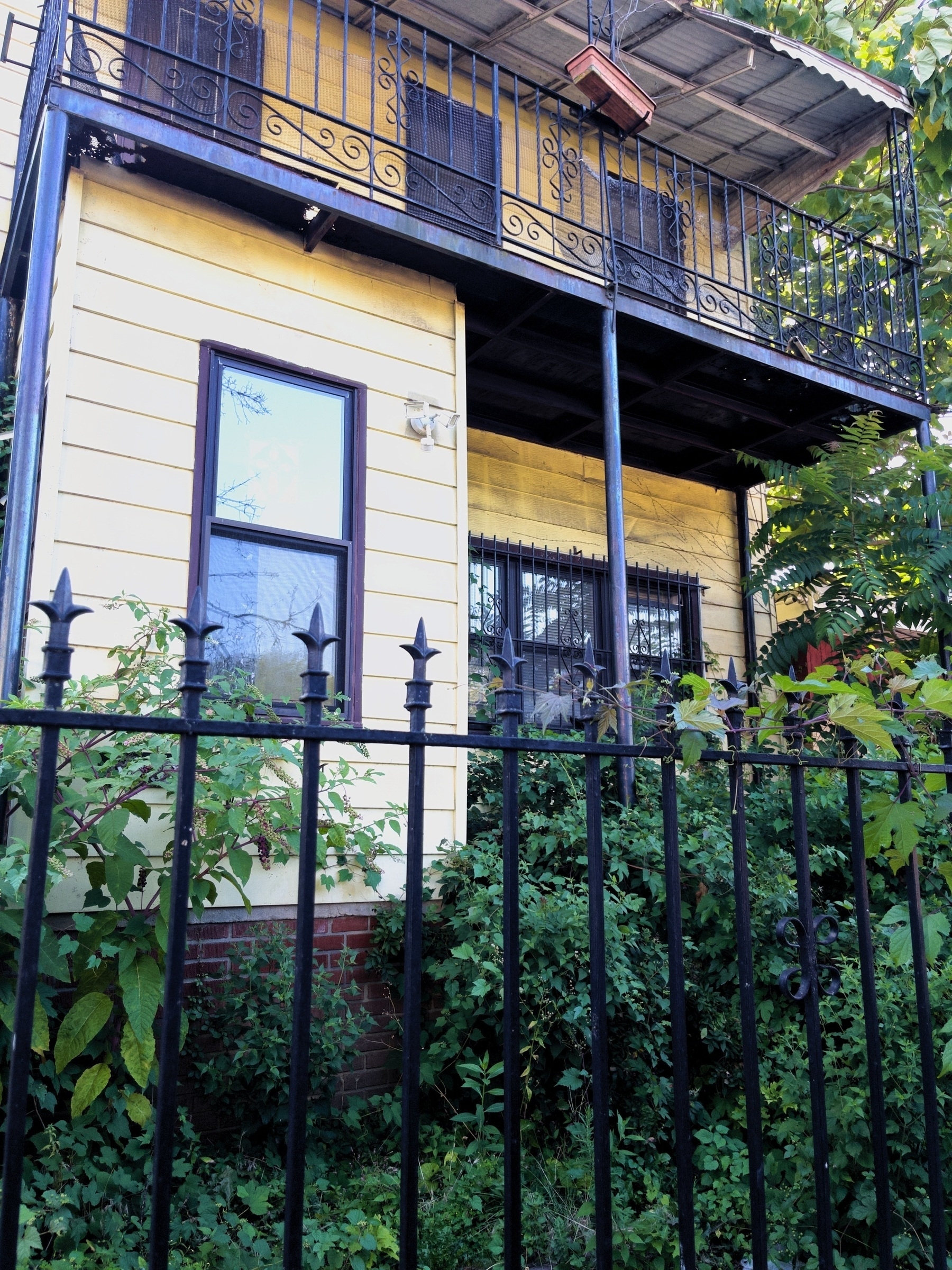 A two-story house with a balcony and wrought iron fencing is surrounded by greenery.