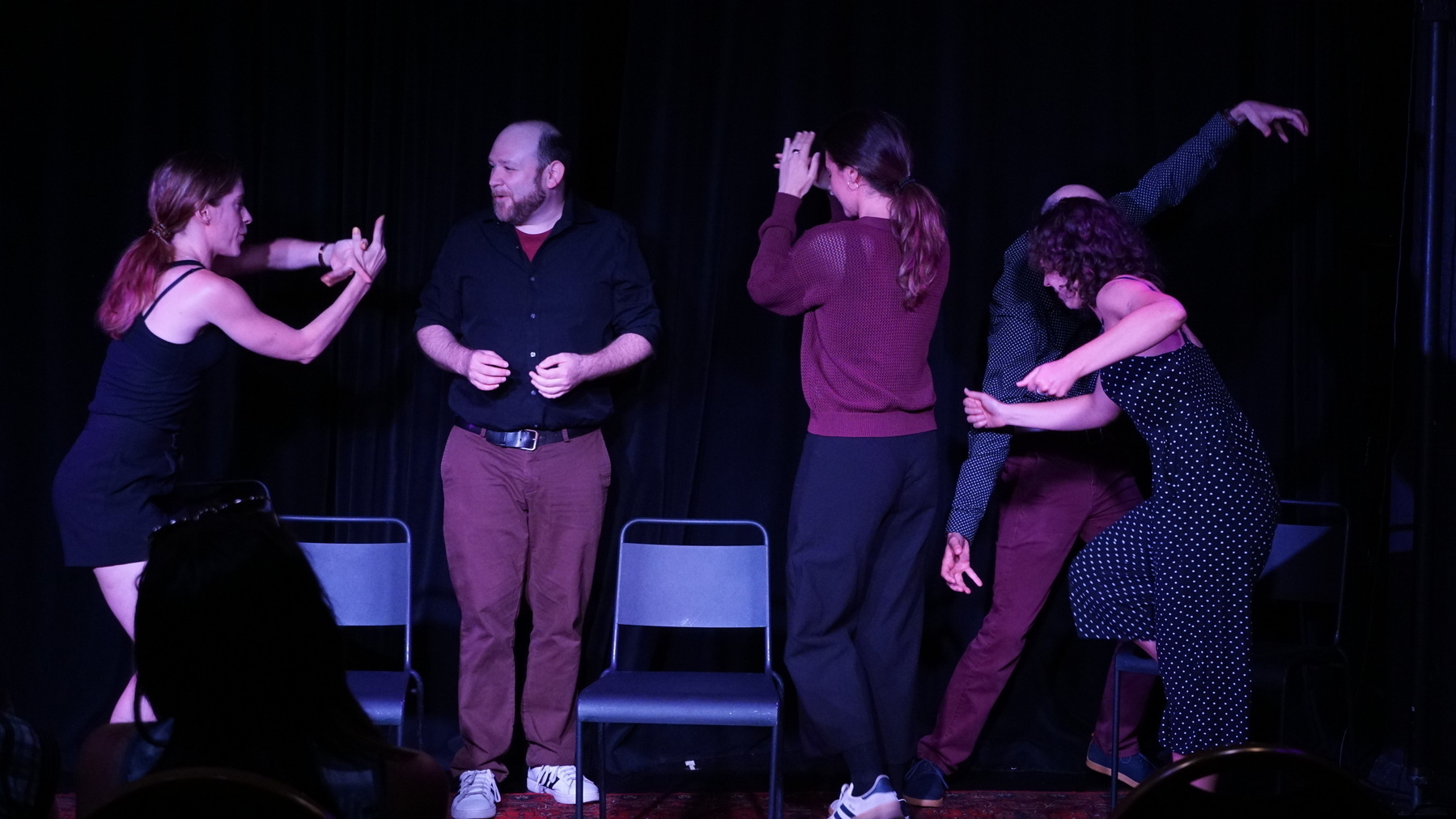 Auto-generated description: Five people are performing an improv scene on stage with expressive gestures and varying stances.