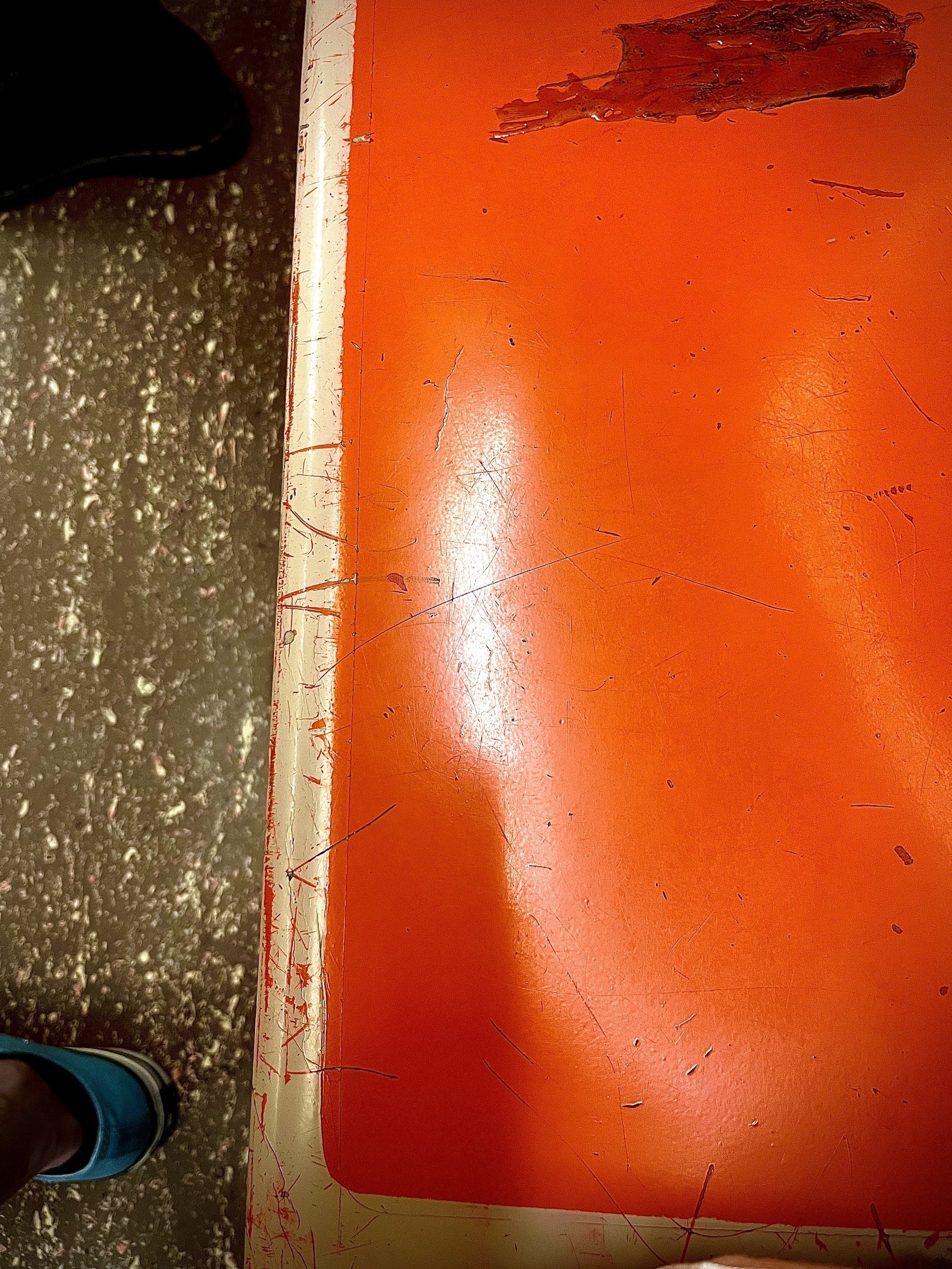 An orange plastic subway seat from above, the brown speckled vinyl flood below, creating a hard line down the middle of the frame.