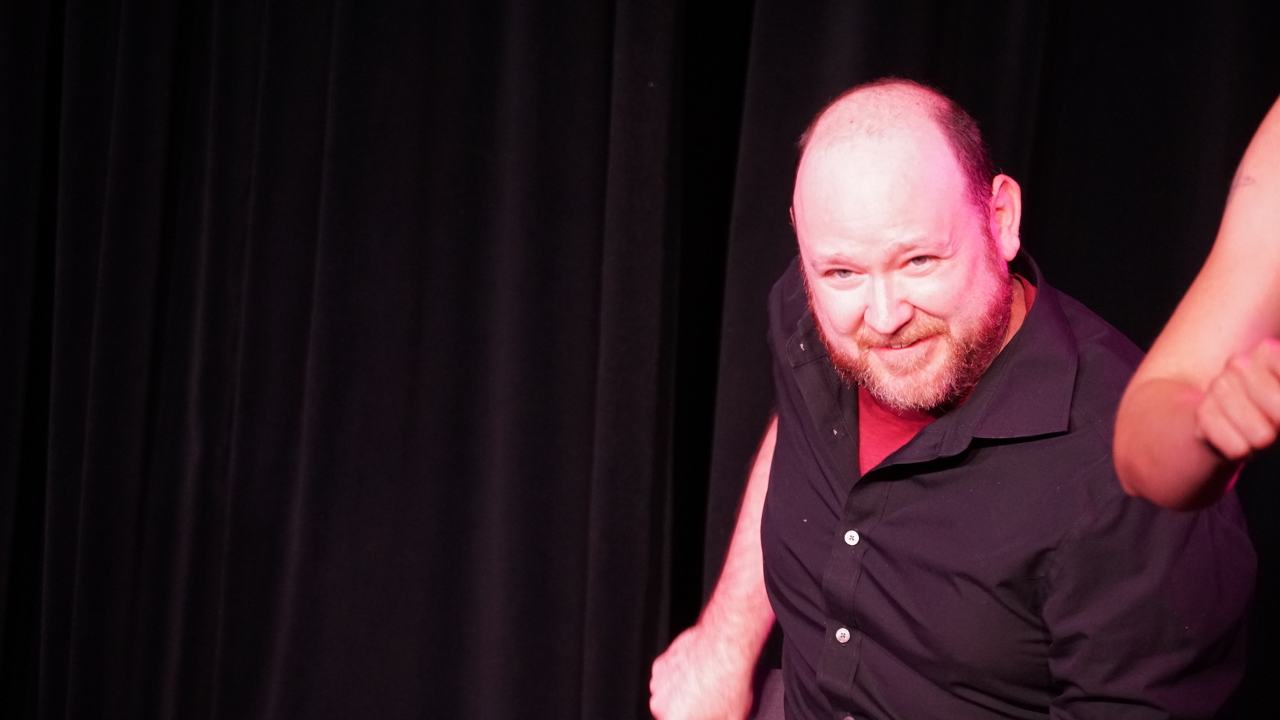 Auto-generated description: A man with a beard and a shaved head is smiling on stage against a black curtain background.