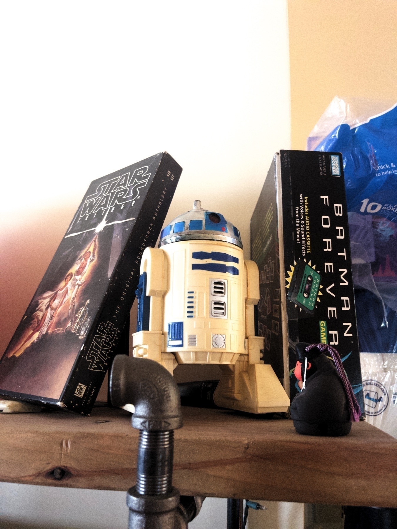 An old R2D2 action figure on top of a wooden shelf. To one side is the Star Wars soundtrack on CD, in a cardboard longbox. It has the original Star Wars poster art. To the other side is the Batman Forever audio cassette board game.