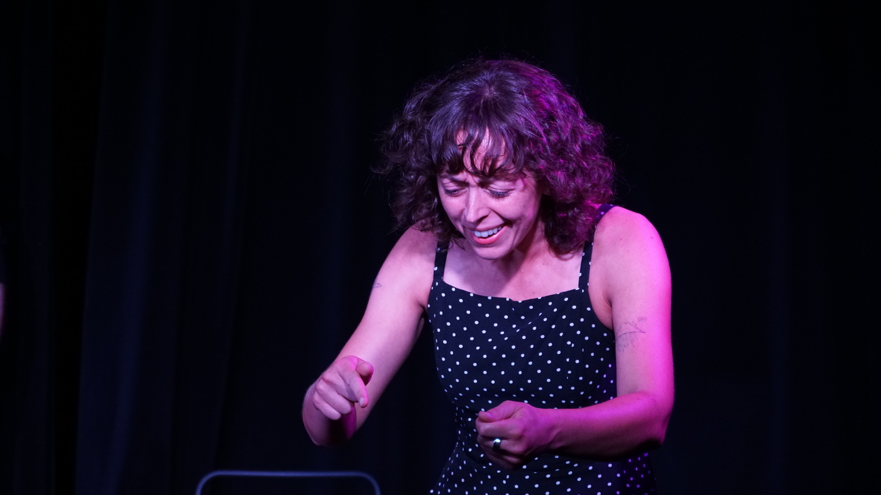 Auto-generated description: A woman with curly hair, wearing a polka dot dress, is performing energetically on stage with a dark background.