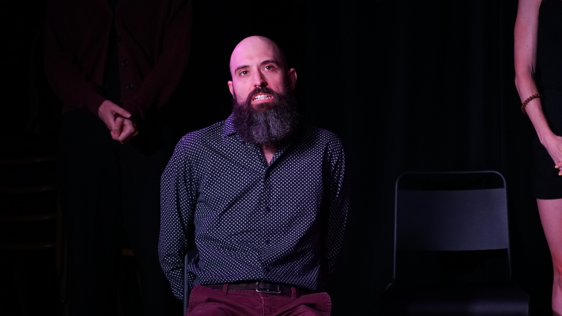 Auto-generated description: A bearded man with a patterned shirt sits on a chair against a dark background, hands behind his back.