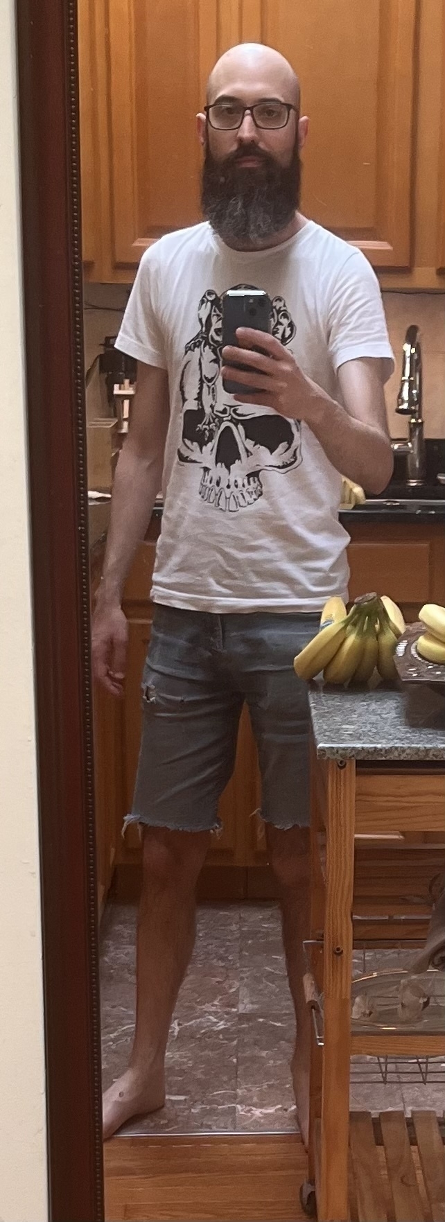Auto-generated description: A bearded man in glasses takes a mirror selfie in a kitchen, wearing a graphic t-shirt and denim shorts, with a cart of bananas nearby.