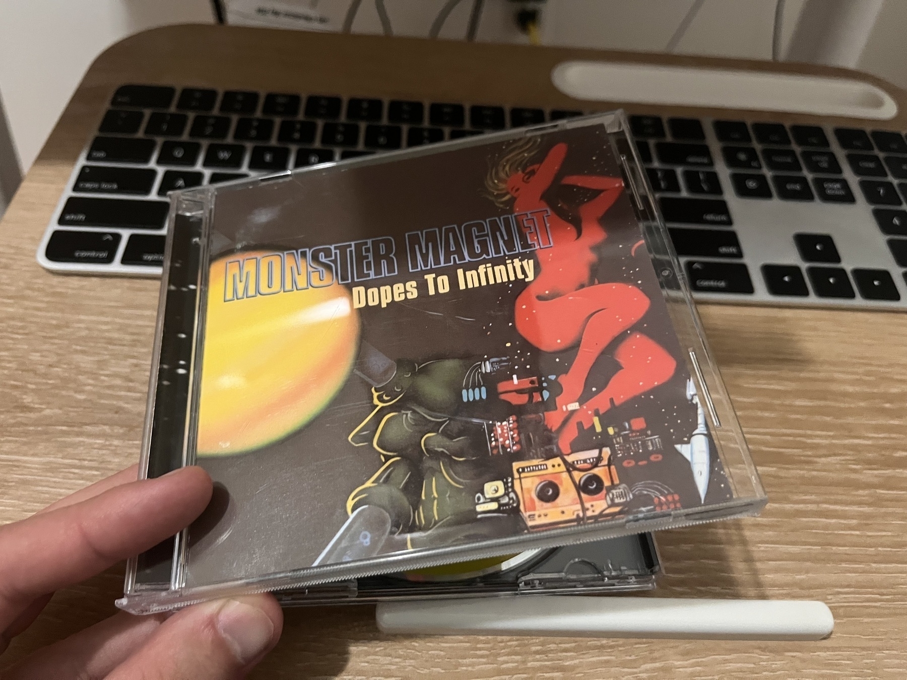 Auto-generated description: A hand holds the Monster Magnet: Dopes to Infinity CD case in front of a keyboard, featuring a colorful sci-fi themed cover.