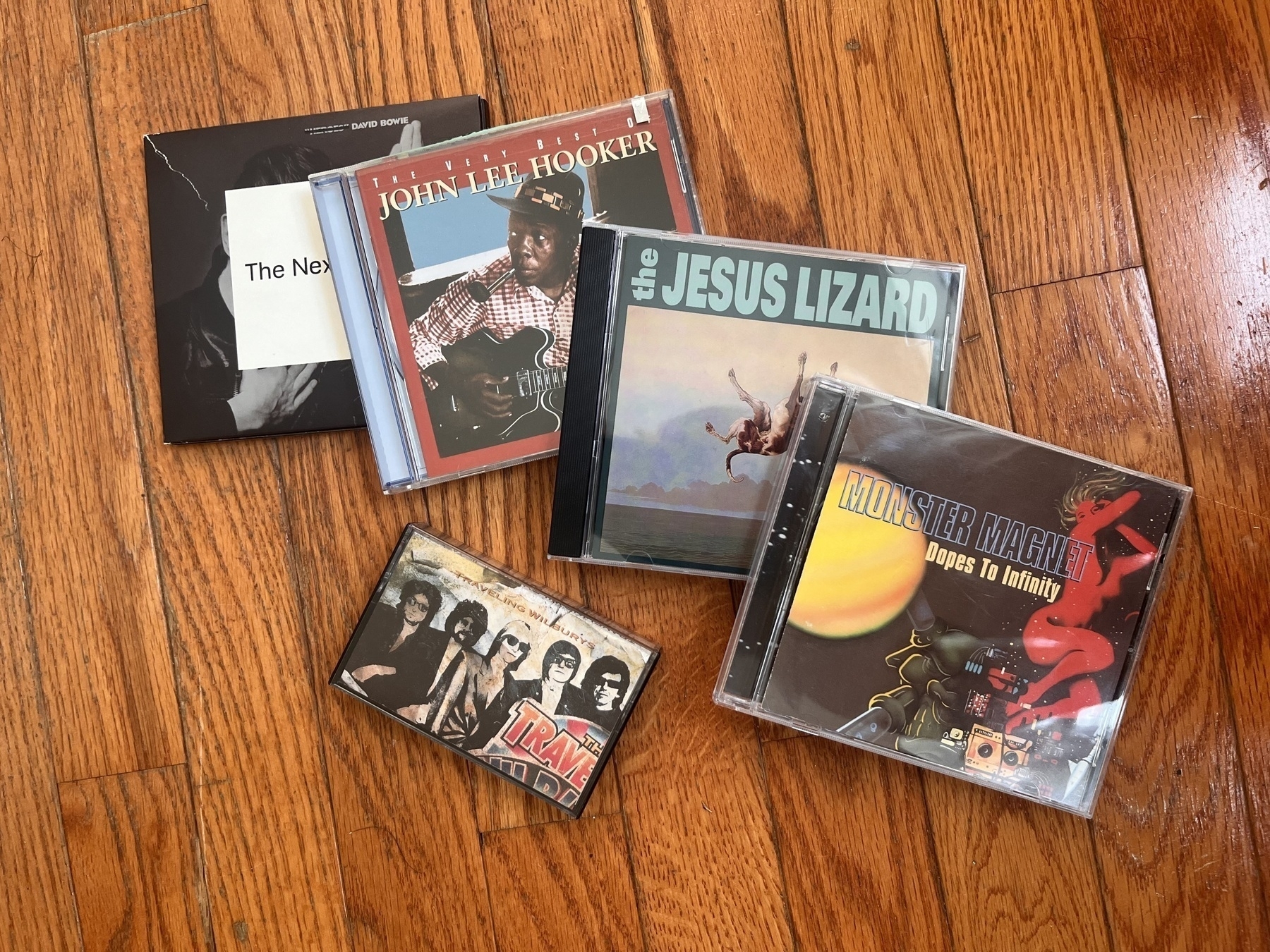 A collection of music CDs and a cassette tape, lying on a wood floor. The CDs are 'The Next Day' by David Bowie, 'The Very Best of John Lee Hooker', 'Down' by The Jesus Lizard, and 'Dopes to Infinity' by Monster Magnet. The cassette is 'The Traveling Wilburys Vol 1'.