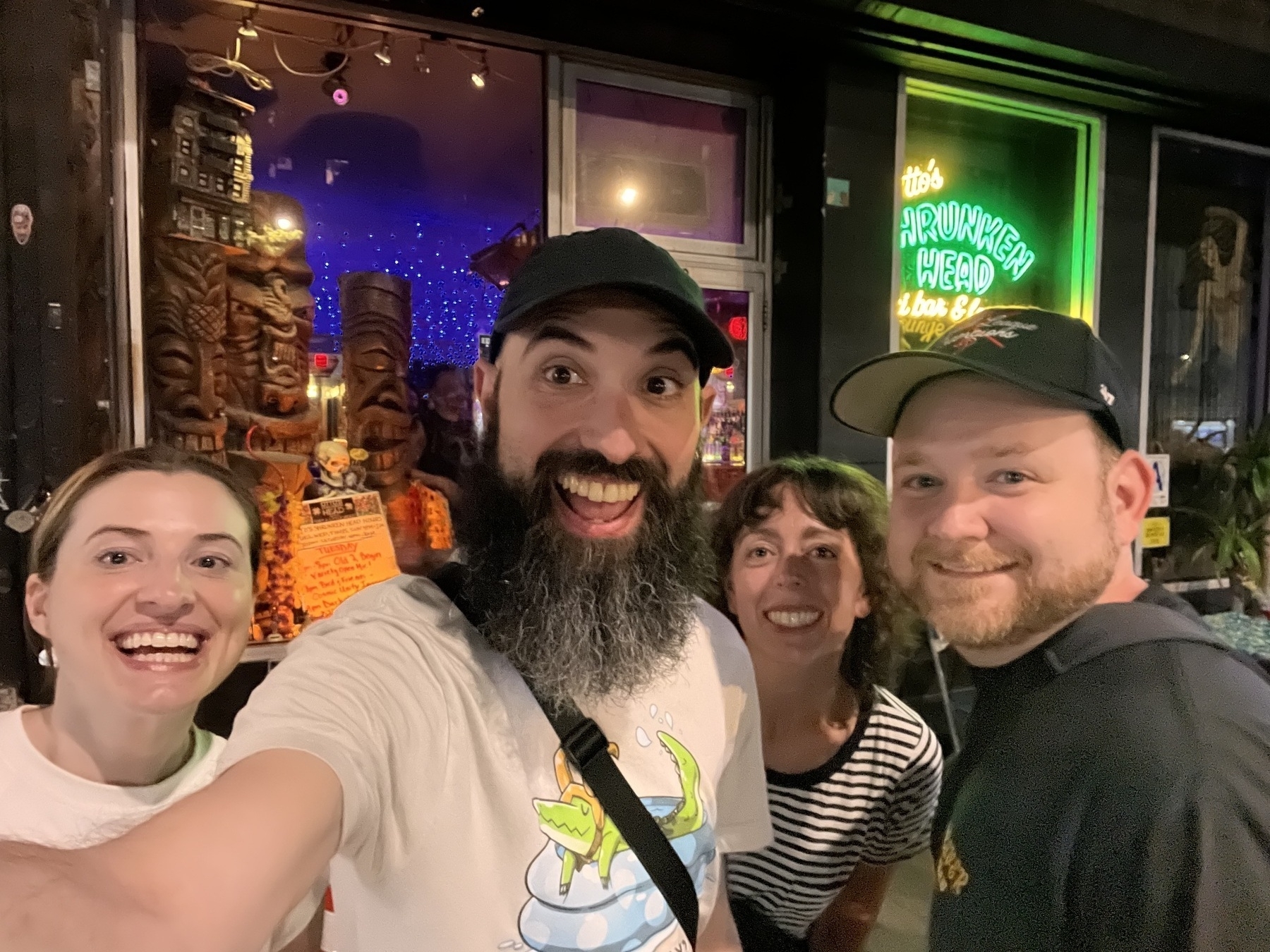 Auto-generated description: Four people are smiling in front of a bar with neon signs and tiki decorations.
