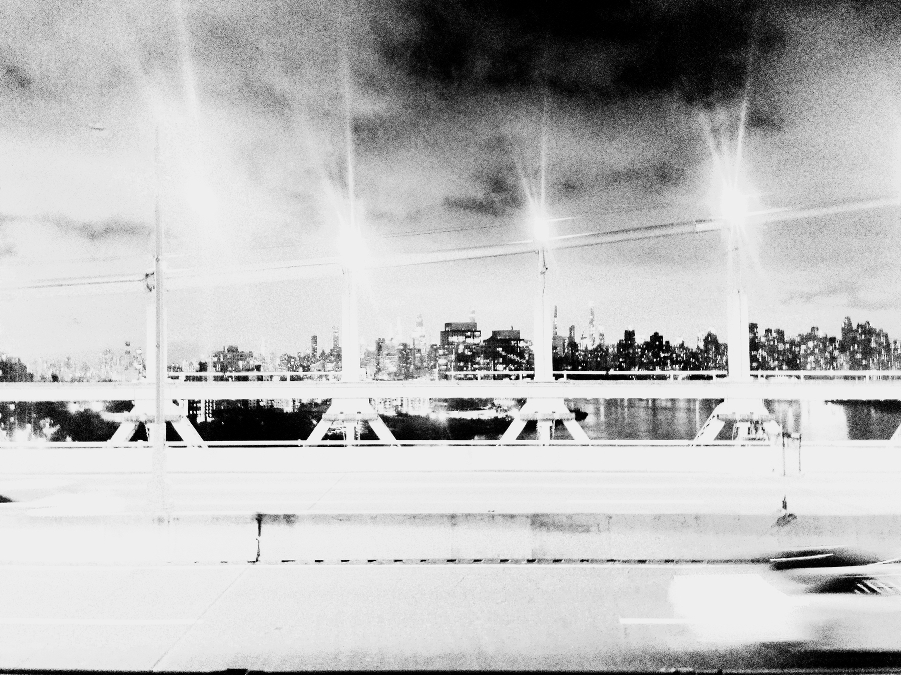 Auto-generated description: A stark black and white photograph captures a blurred vehicle passing over a bridge with a city skyline in the background, illuminated by glowing streetlights.