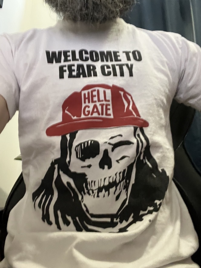 Auto-generated description: A person is wearing a white shirt featuring a graphic of a skeleton with an eye patch, long hair, and a red cap that says HELL GATE with the text WELCOME TO FEAR CITY above the graphic.