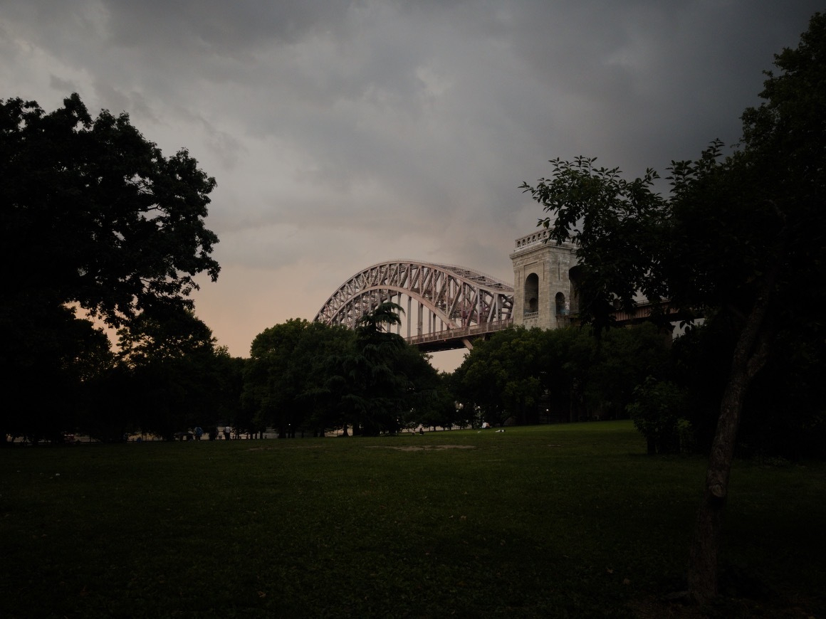 Auto-generated description: A large steel arch bridge spans across a green park area under a cloudy sky during dusk.
