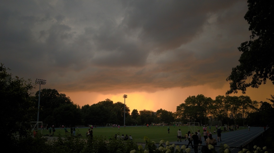 Auto-generated description: A soccer field is surrounded by trees and groups of people under a dramatic sunset sky with dark, heavy clouds.